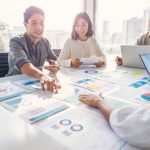 Multi racial diverse group of people working with Paperwork on a board room table at a business presentation or seminar. The documents have financial or marketing figures, graphs and charts on them. People are pointing to different documents. There are laptops and digital tablets on the table